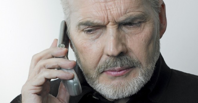 A senior attractive caucasian white man with grey short hair, beard and serious thinking expression holding a mobile phone in his hand, listening, looking down.